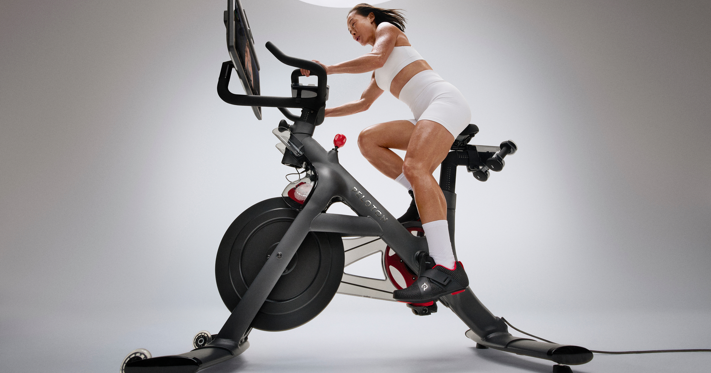 A woman riding a Peloton bike with the touchscreen angled toward her against a grey studio background